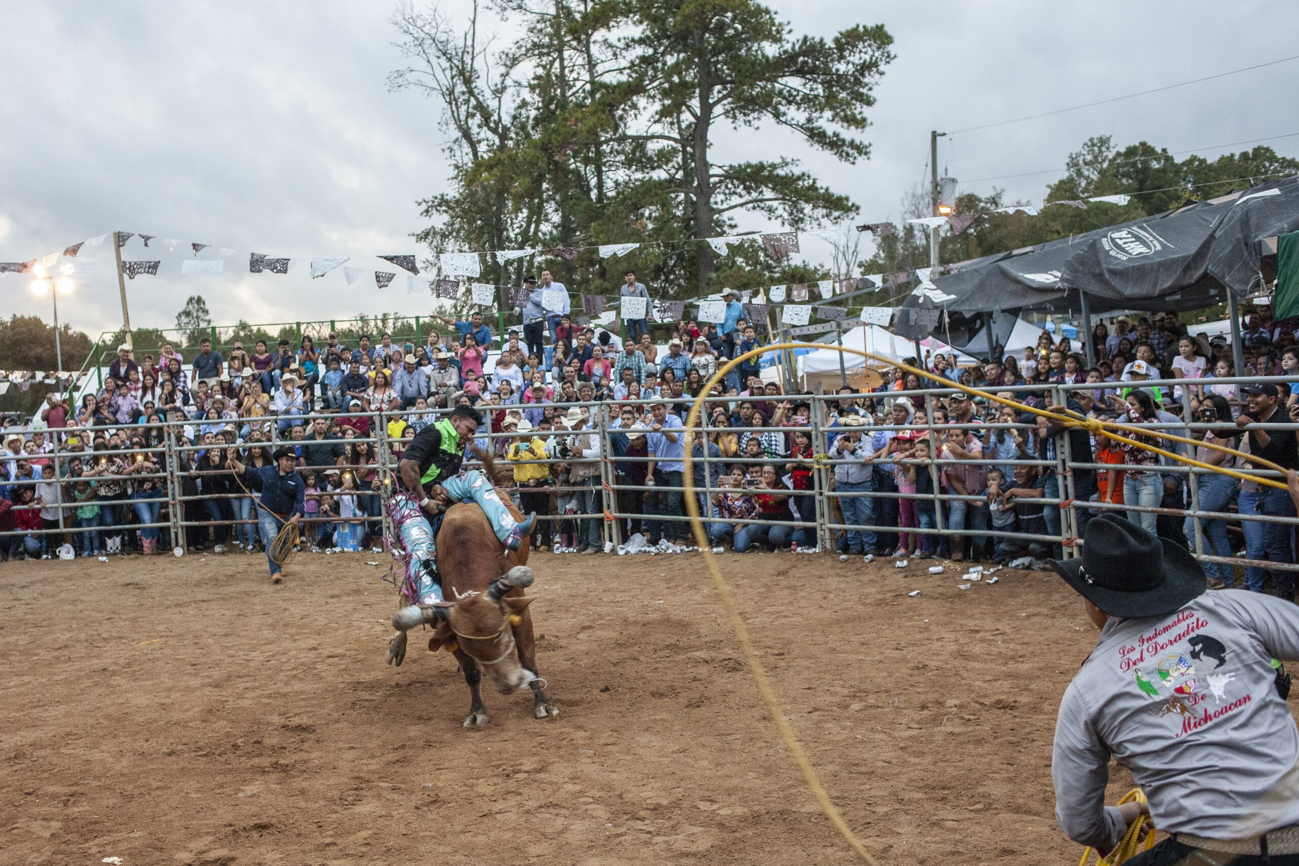 A person struggles to sit on a bull while two people in cowboy hats stand on either side of the bull, swinging lassos. They are located inside a dirt-covered ring as a crowd looks on from outside a fence.