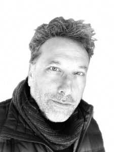  Rodrigo Dorfman, a person with short hair and a stubble beard, is pictured in black and white, wearing a scarf, a black jacket and gazing into the camera behind a white backdrop.