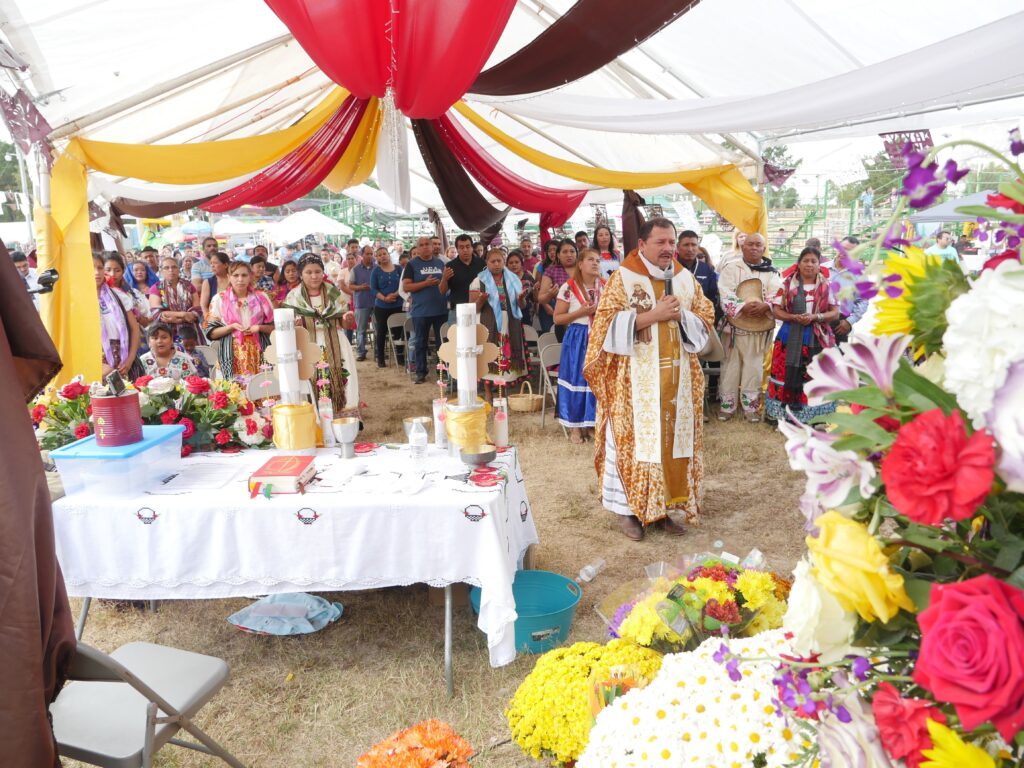 Padre León, wearing a long white and brown religious gown, speaks into a microphone while a large crowd of people in colorful attire stand behind him under a white tent, decorated with banners and bouquets of flowers.