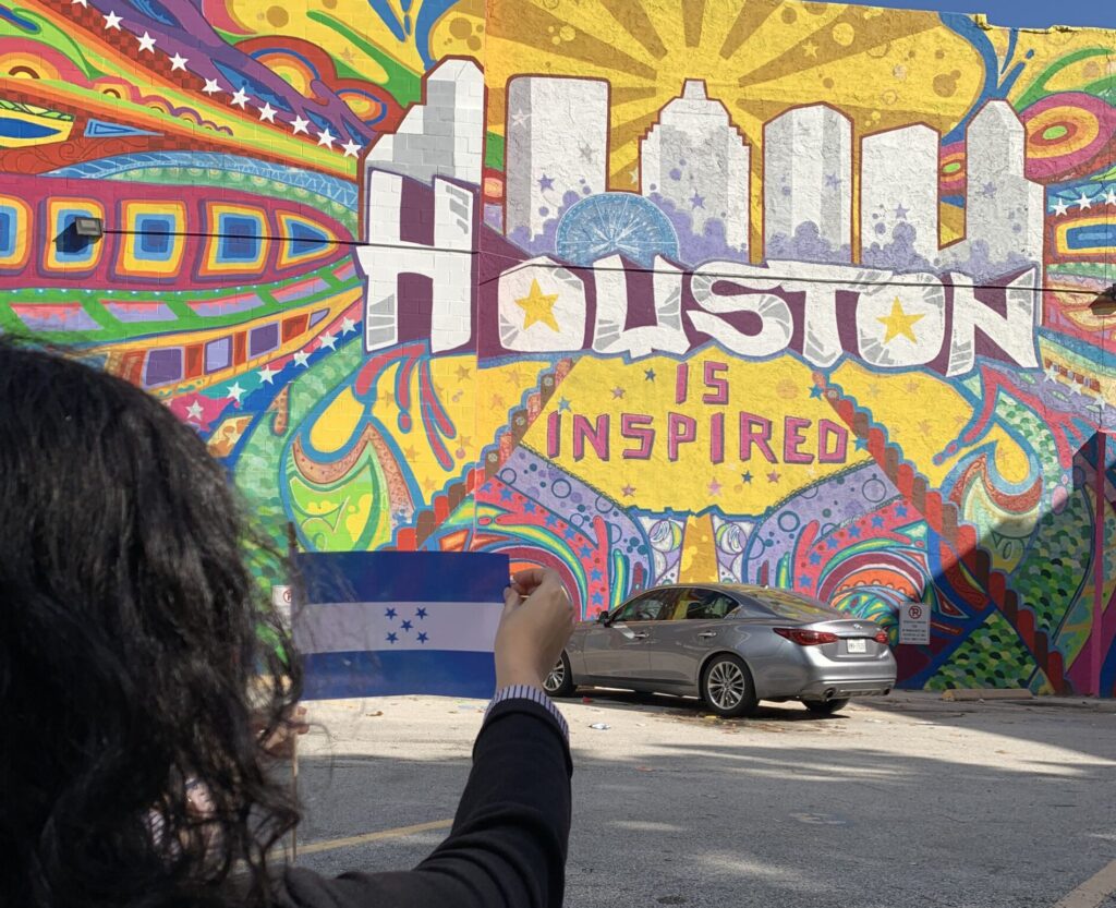 photograph of a woman holding a Honduran flag in front of a mural that reads "houston is inspired" 